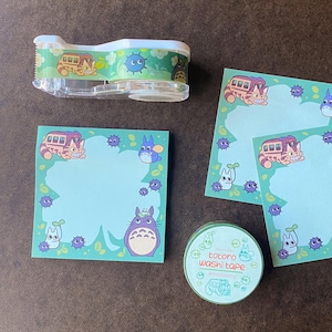 May include: A clear tape dispenser, sticky notes, and washi tape featuring characters from the animated film "My Neighbor Totoro." The items have a light blue and green color scheme with illustrations of the Catbus and Totoro.