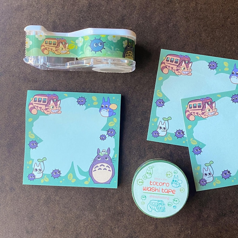 May include: A clear tape dispenser, sticky notes, and washi tape featuring characters from the animated film "My Neighbor Totoro." The items have a light blue and green color scheme with illustrations of the Catbus and Totoro.