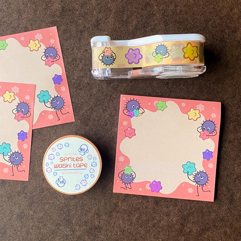 May include: A collection of stationery items featuring a whimsical design. Includes sticky notes, washi tape, and a tape dispenser. The items are decorated with cartoon sprites and colorful floral shapes on a pink background.
