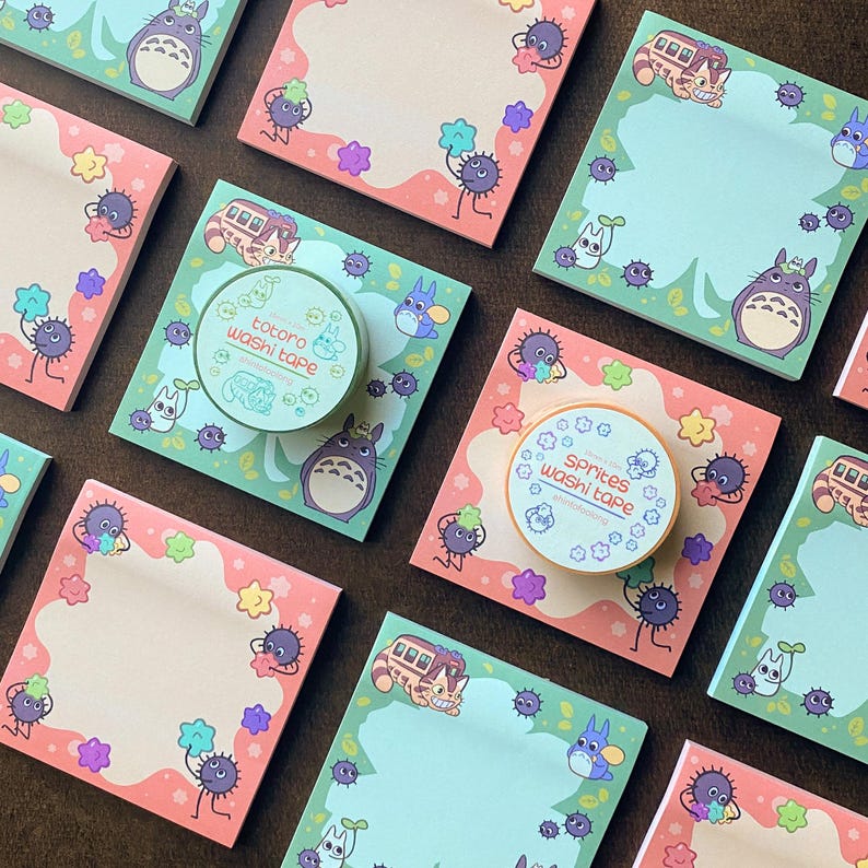 May include: An assortment of stationery items featuring a whimsical design. The image includes square notepads with a pink and green border, and rolls of washi tape. The notepads and tape feature cartoon characters and illustrations.