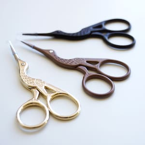 May include: Three pairs of decorative embroidery scissors in a row. The scissors are shaped like birds. One pair is gold, one is brown, and one is black. The scissors are on a white surface.