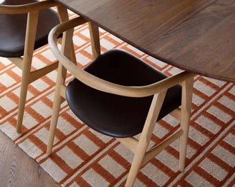May include: A dining room scene featuring a dark wood table with two curved-back chairs. The chairs have brown seats and light wood frames. A patterned rug in orange and cream tones sits beneath the table and chairs. A white bowl filled with fruit is on the table.