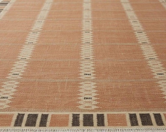 May include: A rectangular rug with a peach-colored background and vertical cream-colored stripes. The rug has a border with alternating brown and peach-colored rectangles and a fringe along the bottom edge. The rug is made of a woven material.