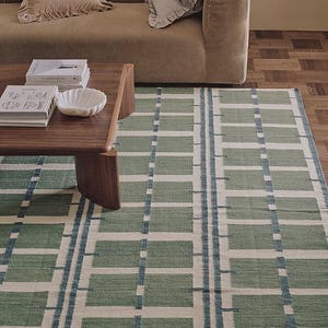 Könnte beinhalten: Rechteckiger Teppich mit geometrischem Muster in Grün, Weiß und Blau. Der Teppich liegt auf einem Holzboden, im Hintergrund ein braunes Sofa und ein Couchtisch. Das Design des Teppichs zeigt rechteckige Blöcke.