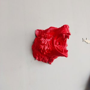 Può includere: Una scultura in 3D stampata in rosso di una testa d'orso con la bocca aperta in un ruggito. La scultura è montata su un muro.