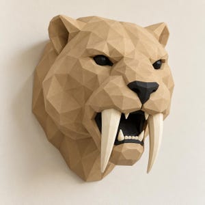 Puede incluir: Una escultura de pared low-poly de una cabeza de tigre dientes de sable. La pieza está construida con material texturizado de color tostado con una superficie geométrica facetada. Presenta colmillos blancos largos, nariz negra y ojos oscuros.