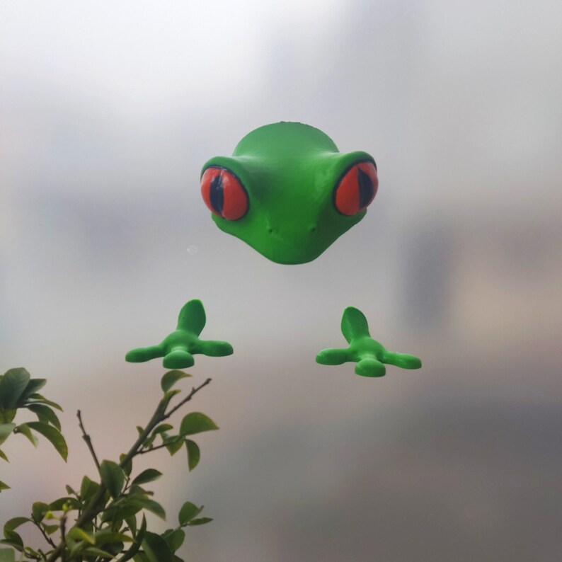 May include: A green frog figurine with red eyes and four green feet. The frog is on a white background.