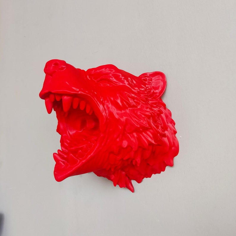 May include: A bright red 3D printed sculpture of a wolf's head with its mouth open in a snarl. The sculpture is detailed with sharp teeth and textured fur.