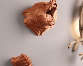 May include: A brown bear head sculpture with an open mouth and sharp teeth, mounted on a wall. A separate brown bear paw sculpture is also mounted on the wall.