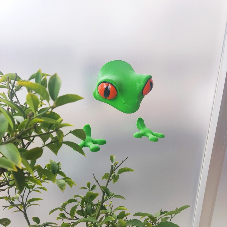 May include: A green frog figurine with large orange eyes and black pupils. The frog is attached to a glass surface with its limbs outstretched.
