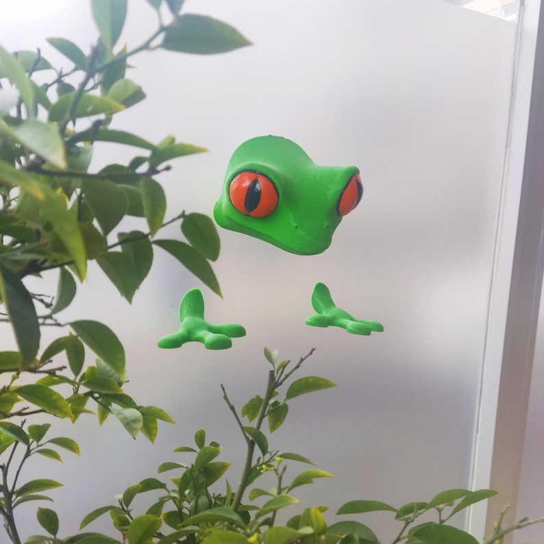 May include: A green frog figurine with large, orange eyes and outstretched arms. The frog is on a white background.