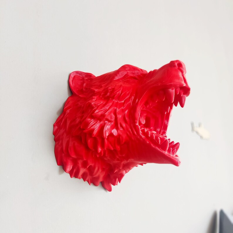 May include: A bright red, 3D printed, wall-mounted sculpture of a wolf's head with its mouth open in a ferocious snarl. The sculpture is detailed with textured fur and sharp teeth.