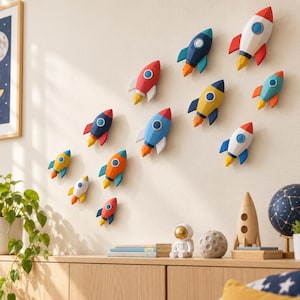May include: A collection of eleven three-dimensional rocket ship wall decals in various colours including red, blue, yellow, orange, and white. Each rocket features a circular window and a pointed nose cone, with stylised flames emerging from the base.