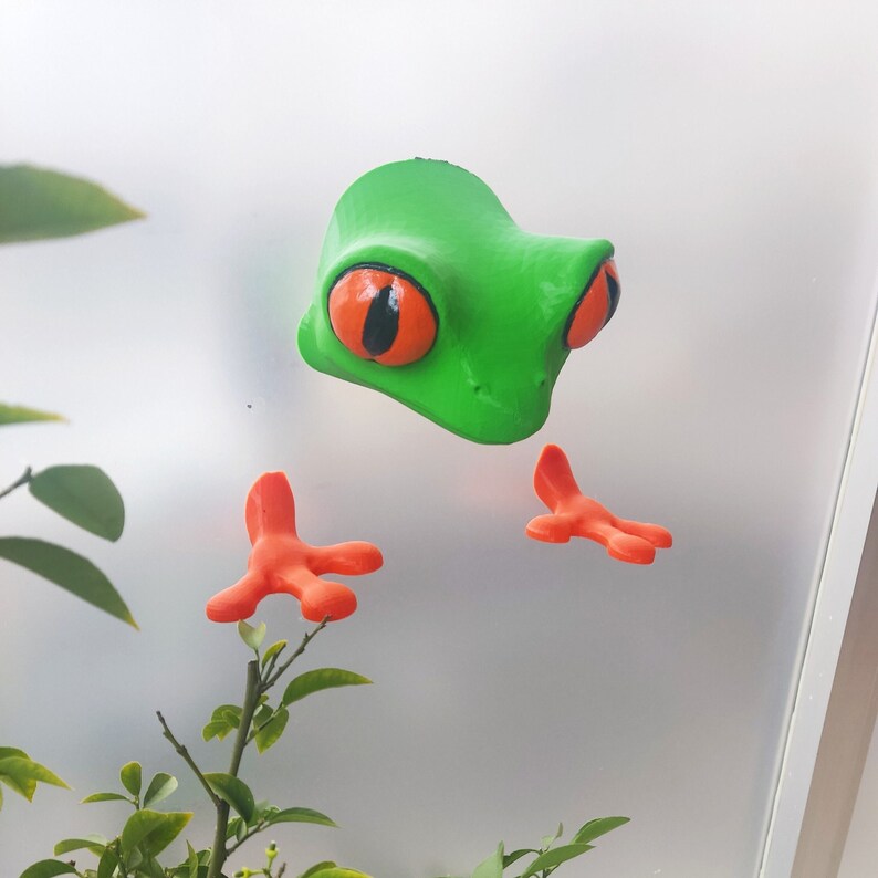 May include: A green frog figurine with orange feet. The frog has large, black eyes with orange pupils.