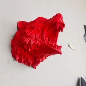 May include: A bright red, 3D printed sculpture of a bear's head with its mouth open in a roar. The sculpture is mounted on a white wall.