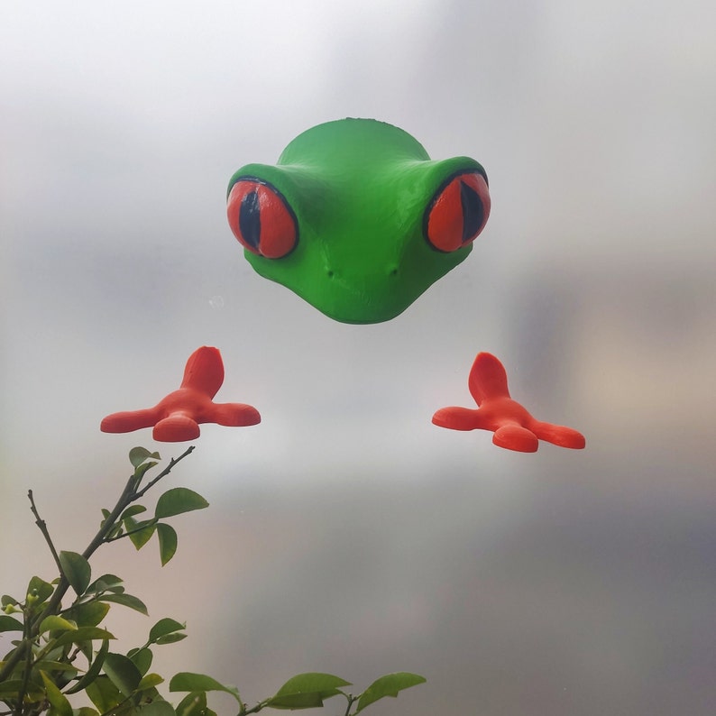 May include: A green frog figurine with red eyes and orange feet. The frog is facing forward with its mouth closed. The figurine is on a white background.