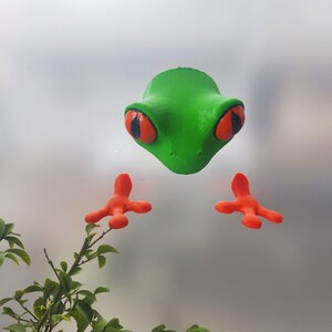 May include: A green frog figurine with orange feet and large red eyes. The frog is facing forward and has a textured surface.