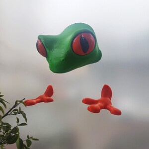 May include: A green frog figurine with large red eyes and orange feet. The frog is posed with its legs outstretched.