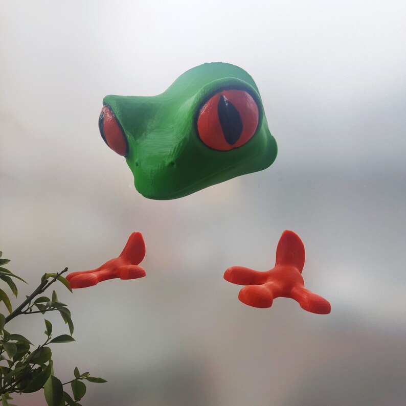 May include: A green frog figurine with large red eyes and orange feet. The frog is posed with its legs outstretched.