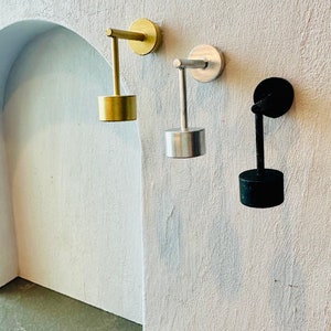 Peut inclure: Trois appliques murales minimalistes en or, argent et noir. Chaque applique comprend une base circulaire et un abat-jour cylindrique, reliés par un bras droit à une fixation murale circulaire. Les appliques sont montées sur un mur blanc texturé.