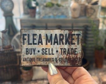 Puede incluir: Un letrero rectangular de metal con un acabado oxidado y desgastado que presenta el texto negro FLEA MARKET, BUY SELL TRADE y ANTIQUE TREASURES & COLLECTIBLES. El letrero tiene una apariencia envejecida con manchas de oxidación naranja y marrón.