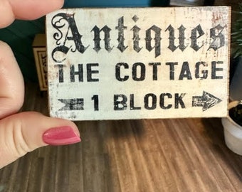 Puede incluir: Un letrero de madera blanco con letras negras que dice "Antiques The Cottage 1 Block" con una flecha negra apuntando hacia la derecha.