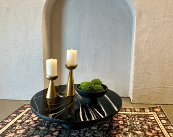 May include: Two gold candle holders with white candles on a black marble table with a small bowl of green moss. The table is on a patterned rug.