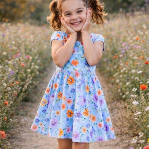 Puede incluir: Un vestido azul claro con un estampado floral vibrante de flores naranjas y moradas, mangas cortas, corpiño ajustado y falda acampanada. La prenda la lleva un niño con el pelo en coletas, sonriendo con las manos sobre las mejillas.