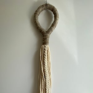 Könnte beinhalten: Ein beigefarbener Makramee-Wandbehang mit einem braunen Seilkreis oben und einer langen Quaste aus beigefarbenem Seil.