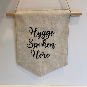 May include: A beige linen banner with the text "Hygge Spoken Here" in black cursive lettering. The banner is hanging from a wooden dowel rod.