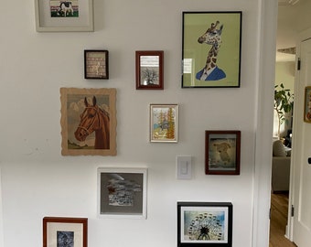 May include: A white wall with a collection of framed pictures, including a giraffe wearing a suit, a horse, a forest scene, and a Ferris wheel.