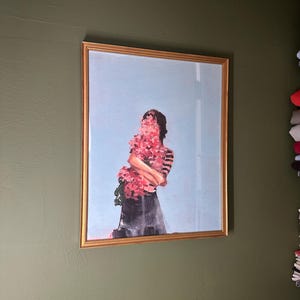 May include: Framed art print featuring a figure obscured by a large bouquet of pink flowers. The figure is wearing a striped top and a dark skirt, set against a light blue sky background. The artwork is encased in a gold frame, mounted on a green wall.