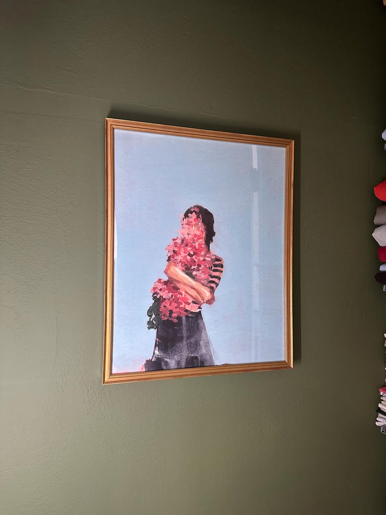 May include: Framed art print featuring a figure obscured by a large bouquet of pink flowers. The figure is wearing a striped top and a dark skirt, set against a light blue sky background. The artwork is encased in a gold frame, mounted on a green wall.