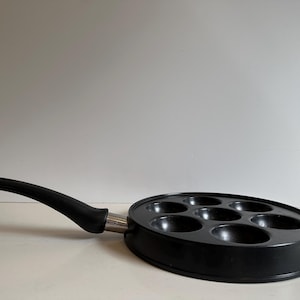 May include: Black, oval-shaped pan with seven circular indentations for cooking. The pan has a long, curved black handle and a silver metal base. The pan is designed for cooking multiple small portions at once.