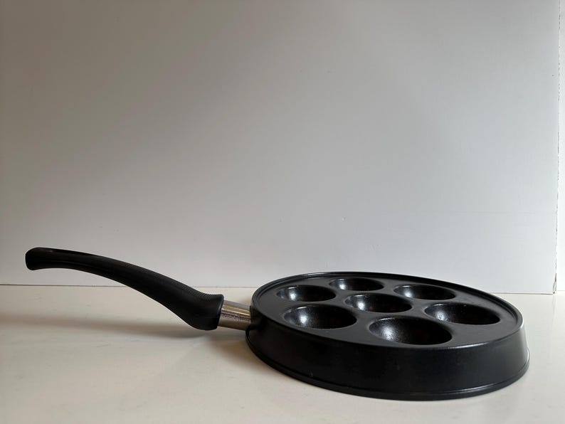 May include: Black, oval-shaped pan with seven circular indentations for cooking. The pan has a long, curved black handle and a silver metal base. The pan is designed for cooking multiple small portions at once.