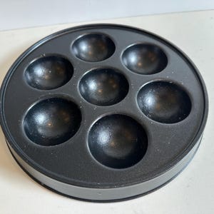 May include: A black, round takoyaki pan with seven spherical molds. The pan has a silver handle with a black grip. The pan is designed for cooking small, ball-shaped Japanese snacks.