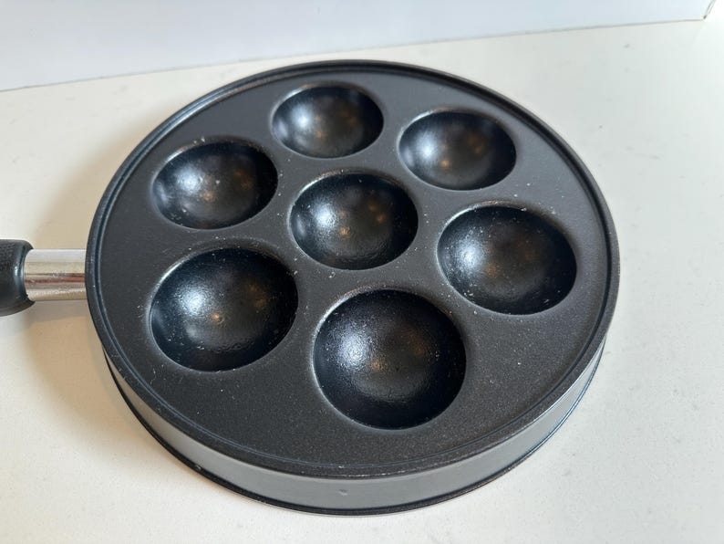May include: A black, round takoyaki pan with seven spherical molds. The pan has a silver handle with a black grip. The pan is designed for cooking small, ball-shaped Japanese snacks.