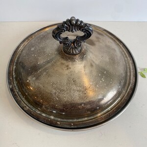 Puede incluir: Plato de servir de plata con tapa y asa adornada. El plato tiene un acabado ligeramente deslustrado.
