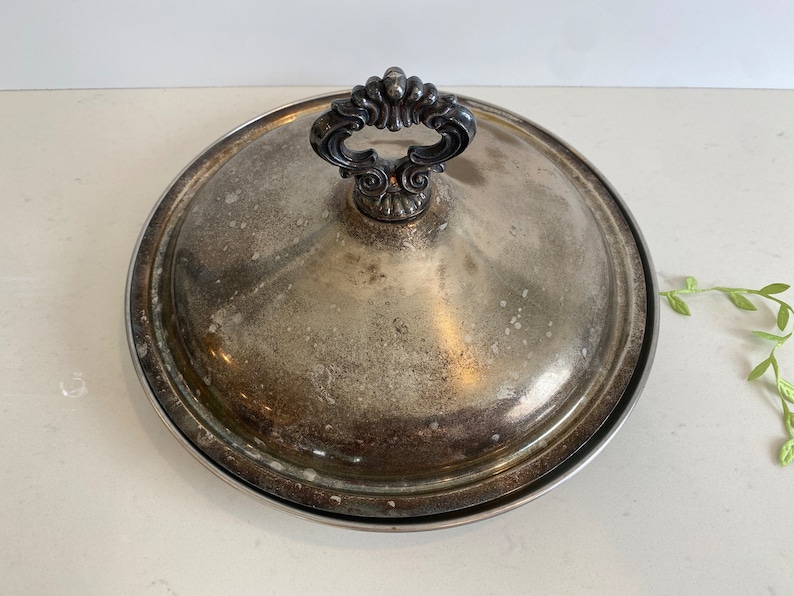 Puede incluir: Plato de servir de plata con tapa y asa adornada. El plato tiene un acabado ligeramente deslustrado.