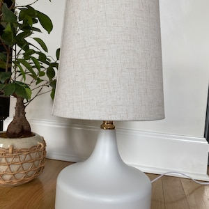 May include: A white ceramic table lamp with a gold accent on the top and a beige linen lampshade.
