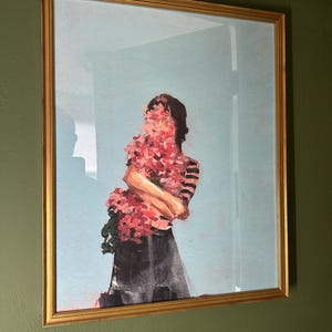 May include: A framed art print featuring a figure holding a bouquet of pink flowers, with the face obscured by the blooms. The figure wears a striped top and a dark skirt, set against a light blue background. The frame is gold.