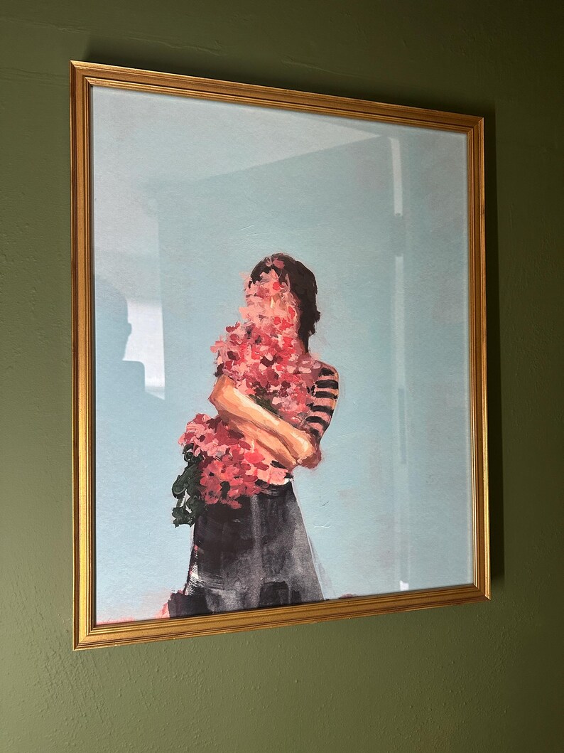 May include: A framed art print featuring a figure holding a bouquet of pink flowers, with the face obscured by the blooms. The figure wears a striped top and a dark skirt, set against a light blue background. The frame is gold.