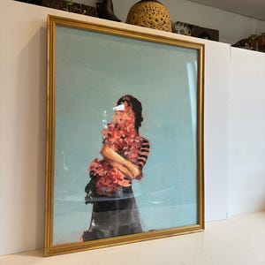 May include: Framed art print featuring a figure holding a bouquet of red flowers against a light blue background. The figure is wearing a black and white striped top and a black skirt. The frame is gold.