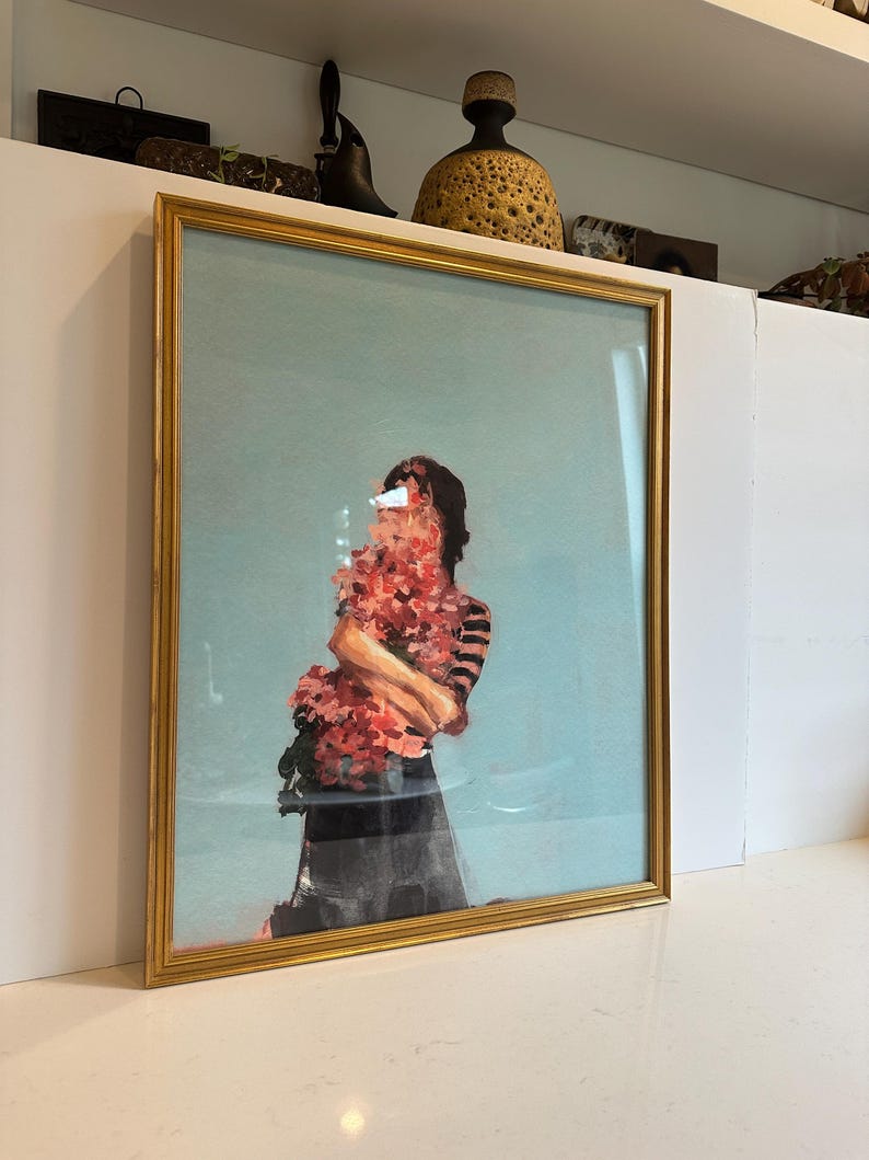 May include: Framed art print featuring a figure holding a bouquet of red flowers against a light blue background. The figure is wearing a black and white striped top and a black skirt. The frame is gold.