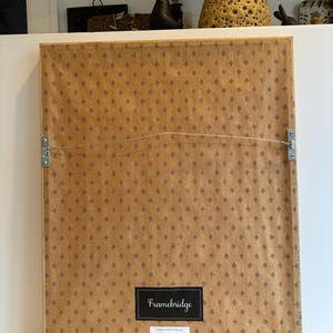 May include: The back of a framed artwork with a brown patterned paper backing. The frame has a wire for hanging and wooden bumpers. A black label reads "Framebridge" with "HANDCRAFTED IN THE USA" below.