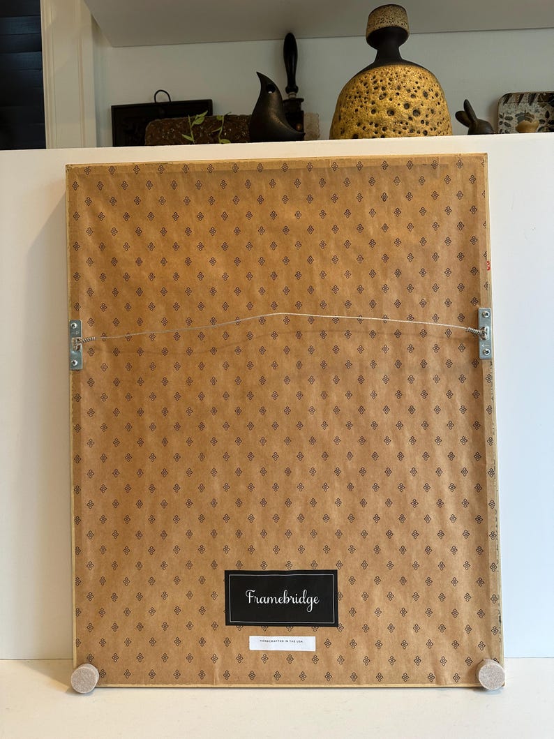 May include: The back of a framed artwork with a brown patterned paper backing. The frame has a wire for hanging and wooden bumpers. A black label reads "Framebridge" with "HANDCRAFTED IN THE USA" below.