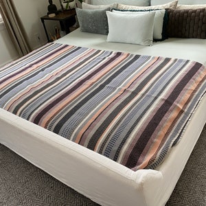 May include: A white bed with a striped throw blanket in shades of pink, gray, and black. The bed has a white headboard and a white footboard.
