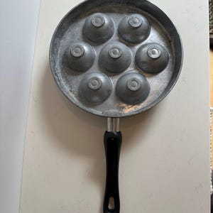 May include: A round, silver-colored pan with seven hemispherical molds. The pan has a black handle. This specialized cookware is designed for making spherical or dome-shaped foods, such as pancakes or eggs.