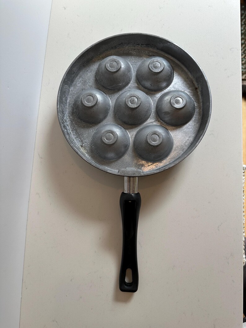 May include: A round, silver-colored pan with seven hemispherical molds. The pan has a black handle. This specialized cookware is designed for making spherical or dome-shaped foods, such as pancakes or eggs.