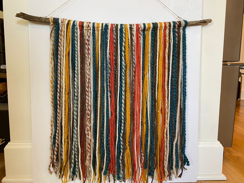 May include: A wall hanging made of yarn in various colors, including teal, brown, white, orange, and yellow. The yarn is tied to a branch and hangs down in long strands. The wall hanging is a bohemian style home decor item.